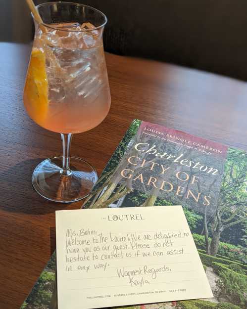 A cocktail beside a welcome note and a magazine about Charleston's gardens on a table.