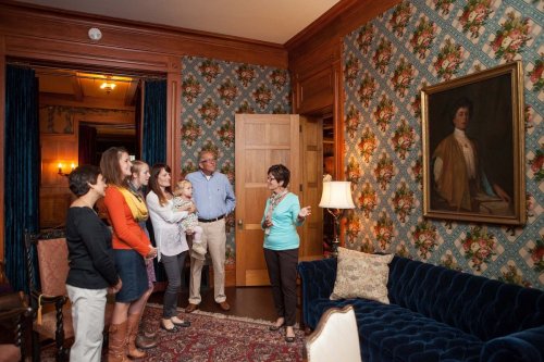 A group of people stands in a room with floral wallpaper, listening to a woman gesturing towards a painting.