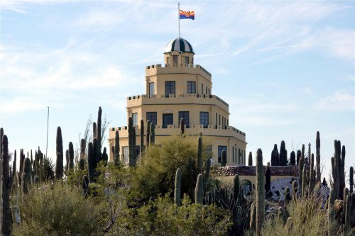 A yellow, multi-tiered building with a dome and flag, surrounded by cacti and desert vegetation under a blue sky.