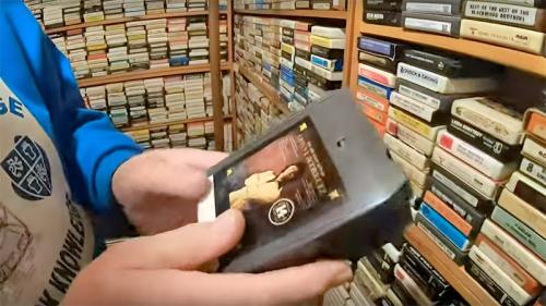 A person holds a VHS tape in front of a shelf filled with various tapes and media.