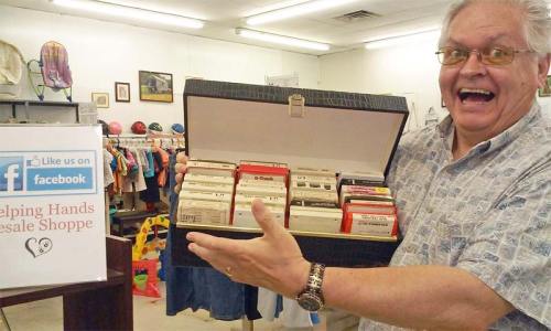 A smiling man holds a case of vintage cassette tapes in a thrift shop, with clothing and items visible in the background.