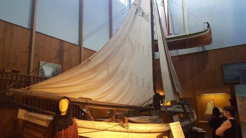 A traditional sailboat displayed in a museum, with wooden walls and other boats visible in the background.