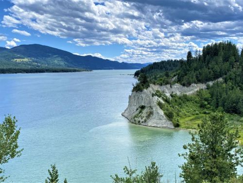 A serene river scene with lush green hills, rocky cliffs, and a cloudy blue sky reflecting on the water.