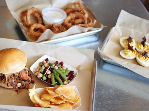 A tray with a pulled pork sandwich, chips, a side salad, onion rings, and deviled eggs on a metal table.