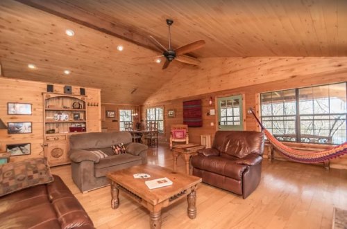 Cozy wooden living room with a ceiling fan, two sofas, a coffee table, and a hammock, featuring large windows.