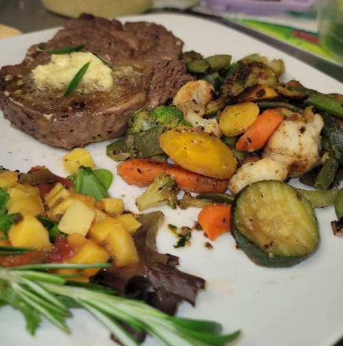 A plate featuring a beef steak with butter, sautéed vegetables, and a mango salsa on a bed of greens.