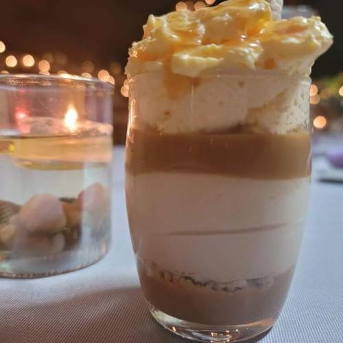 A layered dessert in a glass with cream, caramel, and a crumbly base, beside a candlelit glass of water.
