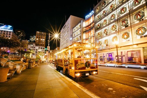 A brightly lit trolley parked on a city street at night, with festive decorations on nearby buildings.