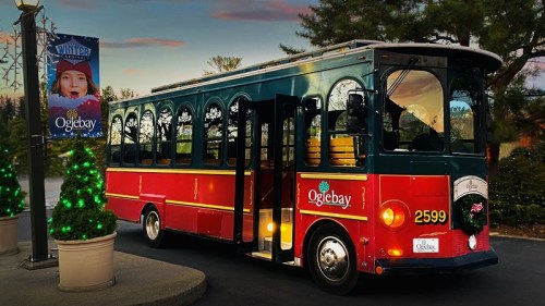 A festive trolley bus parked near a holiday banner, surrounded by trees and decorations at Oglebay.