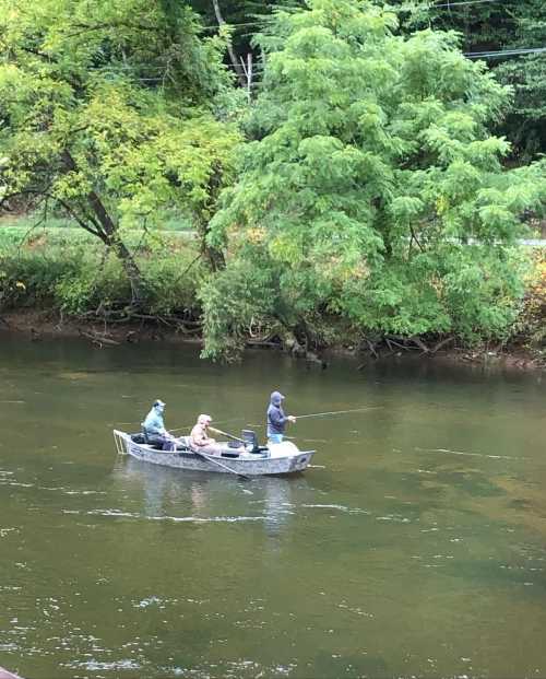 Three people fishing from a small boat on a calm river, surrounded by lush green trees.