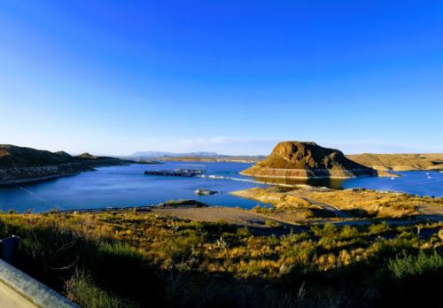 Scenic view of a calm lake surrounded by hills and blue sky, with a rocky island in the center.