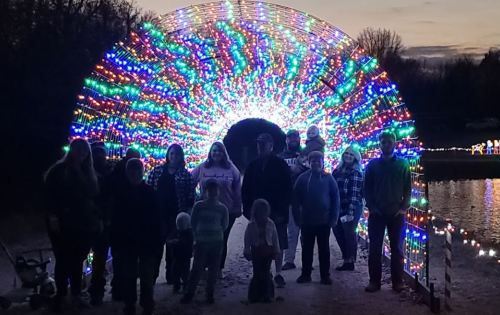 A group of people stands under a colorful, illuminated archway of lights at dusk, with a lake in the background.