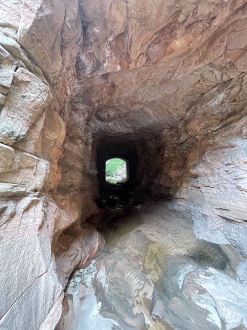 A narrow, rocky tunnel with smooth walls and a small opening at the end, reflecting light and water on the ground.