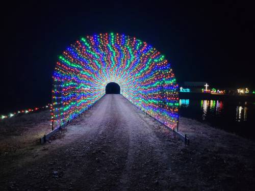 A colorful light tunnel arches over a dirt path, leading to a serene water view at night.
