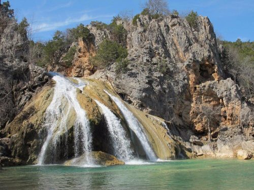 A scenic waterfall cascading over rocky cliffs into a clear blue pool, surrounded by lush greenery and a bright sky.