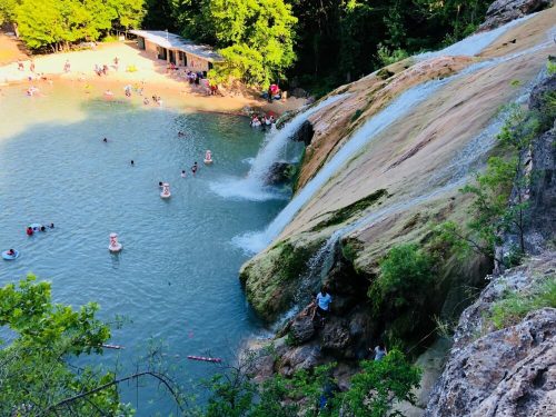 A scenic view of a waterfall cascading into a swimming area, with people enjoying the water and lush greenery surrounding.