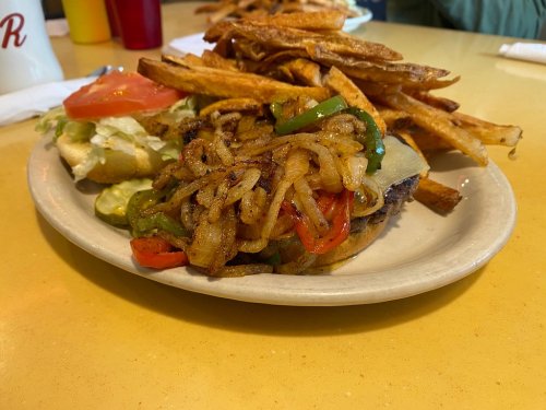 A plate with a burger topped with sautéed onions and peppers, served with lettuce, tomato, pickles, and fries.