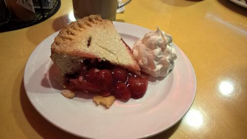 A slice of cherry pie with a flaky crust, topped with whipped cream, on a white plate against a yellow table.