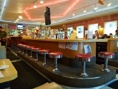 A retro diner interior featuring a long counter with red stools, neon lights, and wooden paneling.
