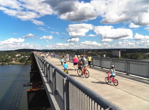 A busy bridge with people walking and children riding bikes, surrounded by blue skies and fluffy clouds.