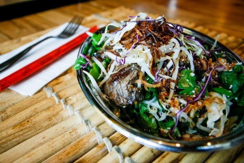A metal bowl filled with a colorful salad topped with shredded cabbage, herbs, and pieces of meat, on a wooden table.
