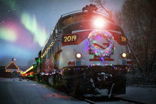 A decorated train with festive lights and a wreath, set against a snowy landscape and colorful northern lights.