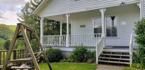 A white house with a porch and swing set in front, surrounded by greenery and hills in the background.