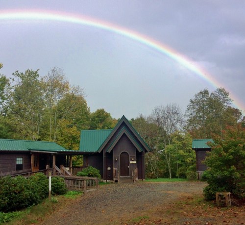 A cabin with a green roof under a rainbow, surrounded by trees and a gravel path.