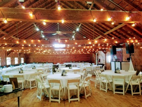 A spacious barn interior with round tables, white chairs, and string lights hanging from the wooden ceiling.