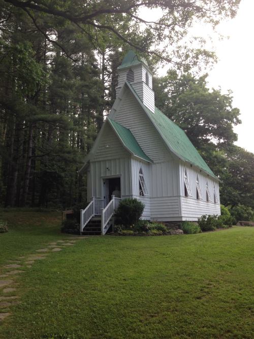 A small white church with a green roof, surrounded by trees and grass, featuring a front porch and steps.