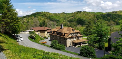 A scenic view of a rustic building surrounded by green hills and trees, with parked cars along a winding road.