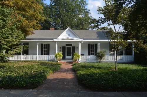 A charming white house with a front porch, surrounded by greenery and trees on a sunny day.