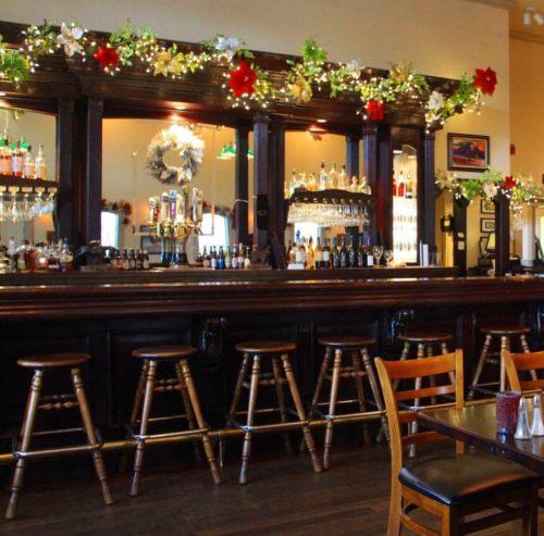 A festive bar decorated with holiday garlands, featuring wooden stools and a well-stocked backlit shelf of drinks.