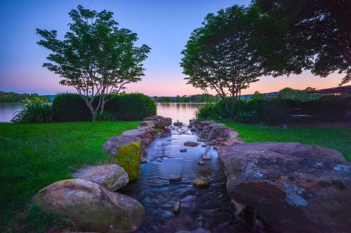 A serene lakeside scene at dusk, featuring a gentle stream flowing over rocks, framed by trees and a calm water surface.
