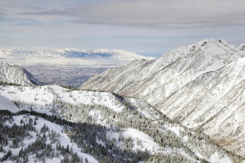 Snow-covered mountains stretch across the horizon under a cloudy sky, with valleys and trees visible below.