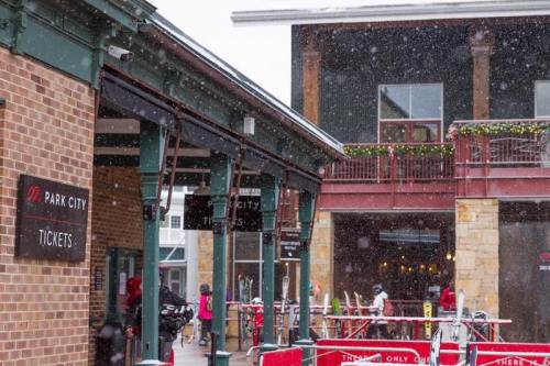 Snow falls over a ski resort ticket area, with people and ski equipment visible in a bustling winter scene.