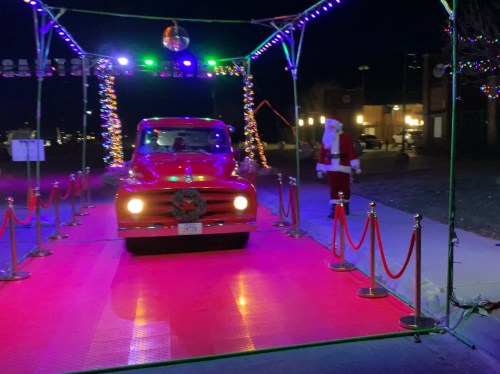 A vintage red truck drives through a festive, illuminated pathway with Santa standing nearby.