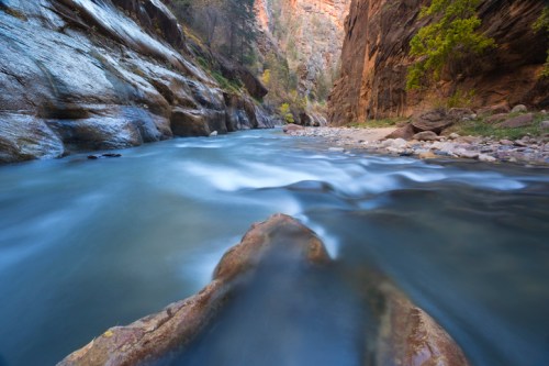 A serene river flows through a narrow canyon, with smooth rocks and towering cliffs on either side.