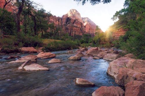 A serene river flows through a lush landscape, with towering red rock formations and sunlight filtering through trees.
