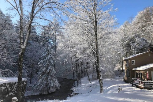 A snowy landscape with frosted trees, a river, and a cozy house under a clear blue sky.