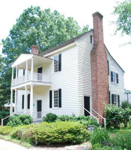 A two-story white house with a porch, brick chimney, and lush greenery surrounding it.