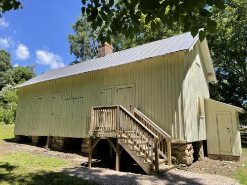 A light green wooden building with a metal roof, featuring a staircase and surrounded by trees and blue sky.