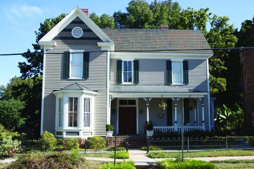 A two-story gray house with white trim, green shutters, and a front porch, surrounded by trees and a garden.
