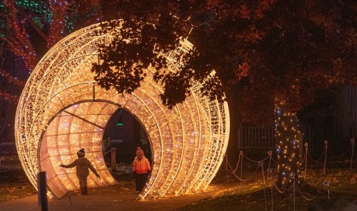 Two people walk through a glowing, spiral light tunnel surrounded by trees adorned with festive lights.