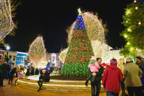 A festive scene with a large Christmas tree adorned with lights, surrounded by people enjoying the holiday atmosphere.
