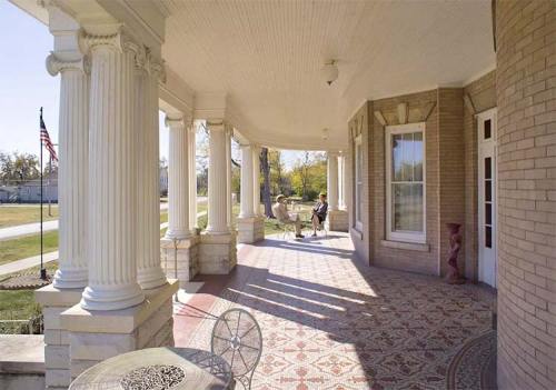 A spacious porch with columns, decorative tiles, and a view of a garden, featuring a person seated in the background.