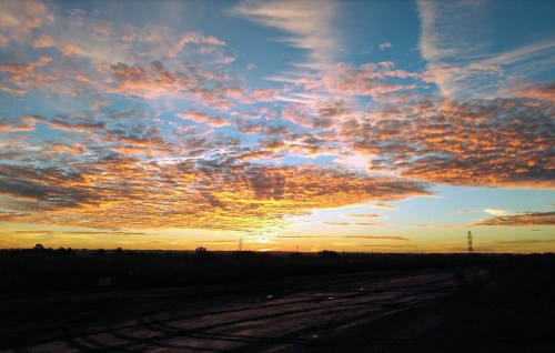 A vibrant sunset with colorful clouds over a rural landscape, featuring a dirt road and distant power lines.
