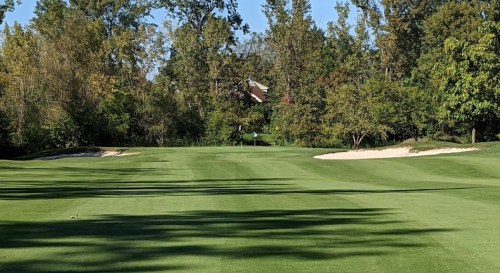 A lush green golf course with trees in the background and a sand trap on the right side. Clear blue sky above.