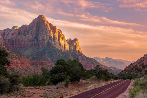 A winding road leads through a scenic landscape of red rock mountains and lush greenery under a colorful sky.
