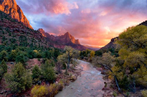 A scenic view of a river winding through a canyon at sunset, with vibrant clouds and colorful rock formations.